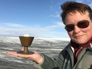 Julie Taylor holding a chalice in her hand. Short brown hair, wearing glasses, smiling in front of snowy outdoor vista.